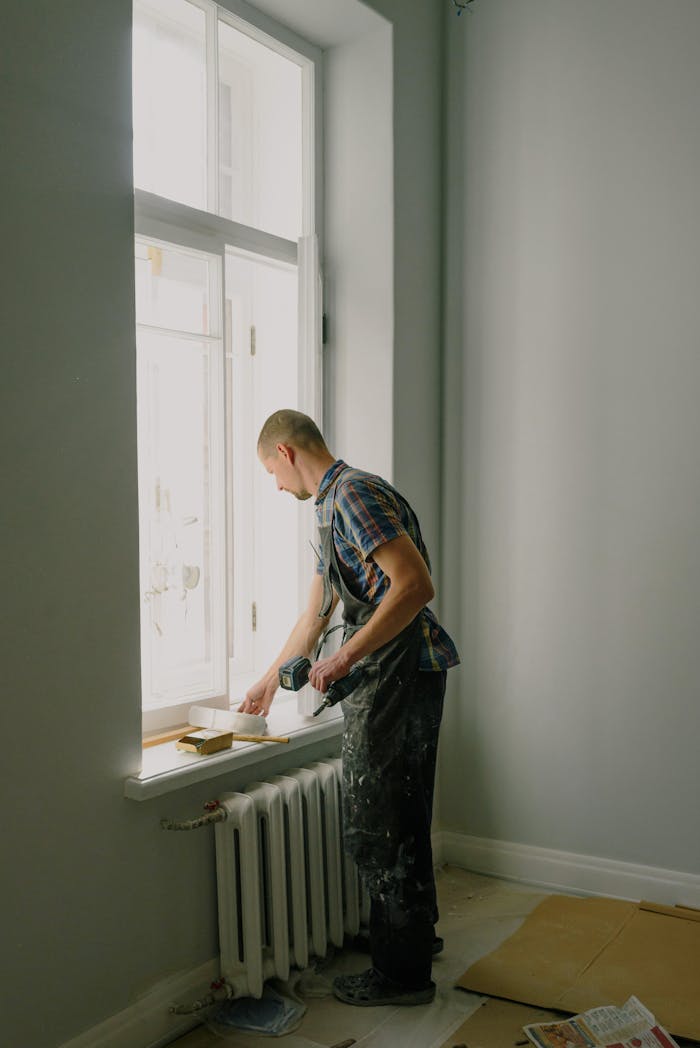 A man in overalls painting a window ledge inside a home, bright daylight.